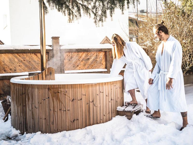 Zwei Personen in Bademänteln betreten einen hölzernen Whirlpool im Schnee.