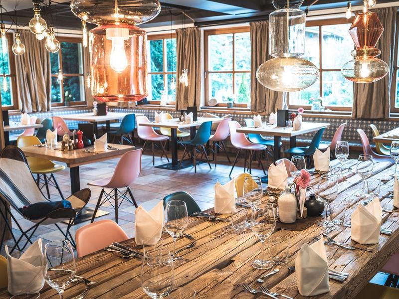 Gemütlicher, rustikaler Restaurantbereich mit Holztischen, bunten Stühlen und großen Fenstern.