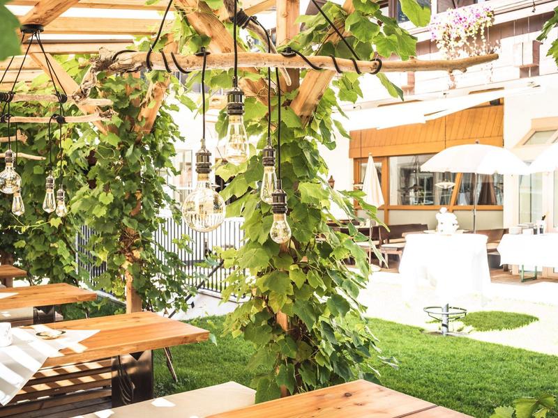 Sonnige Terrasse mit Holztischen, Stühlen und grünen Pflanzen unter einem Holzpergola-Dach.