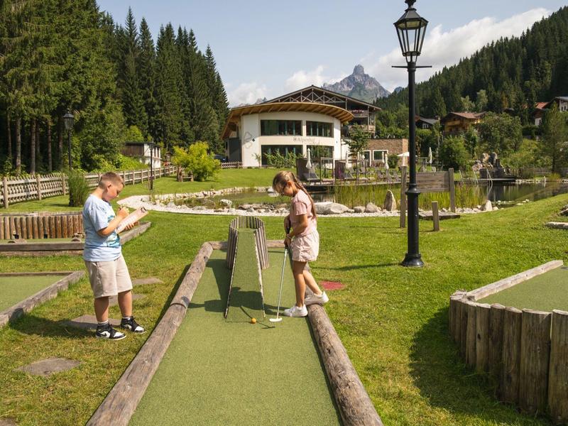Twee personen spelen minigolf op een zonnig clubterrein met bomen en bergen op de achtergrond.