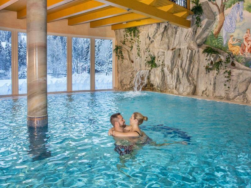 Twee mensen omhelzen elkaar in een binnenzwembad met rotsmuur en grote ramen op de achtergrond.
