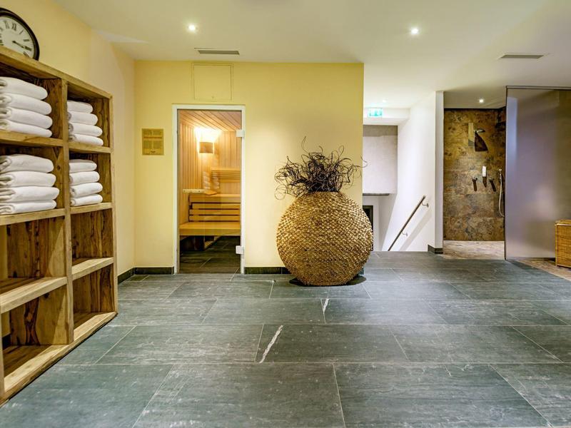 Área de bienestar luminosa con suelo de piedra, estante de madera y gran jarrón decorativo en el centro.