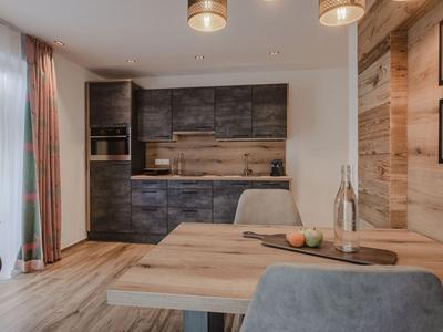 Área de cocina moderna con gabinetes oscuros, mesa de madera y sillas grises en habitación de hotel.