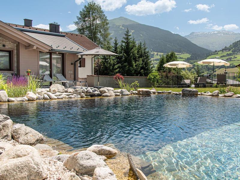 Una casa con una piscina natural y vistas a la montaña bajo un cielo azul con nubes.