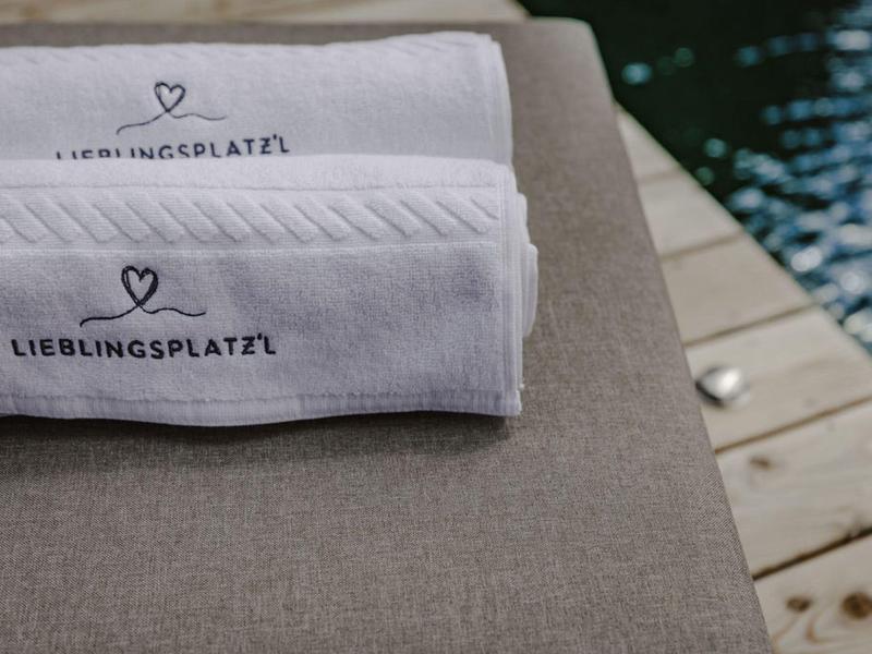 Dos toallas blancas con corazón y texto sobre cojín gris junto a un muelle de madera en el agua