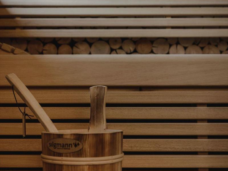 Sauna acogedora con bancos de madera y un cubo con un cucharón frente a leña apilada.