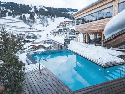 Piscina exterior en invierno con terraza de madera nevada y vista a montañas nevadas y edificios del hotel.