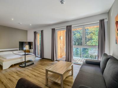 Brightly furnished hotel room with large window, sofa, bed, and wooden floor.