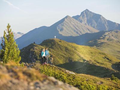 Zwei Wanderer auf einem hügeligen Pfad mit Bergpanorama und blauem Himmel im Hintergrund.