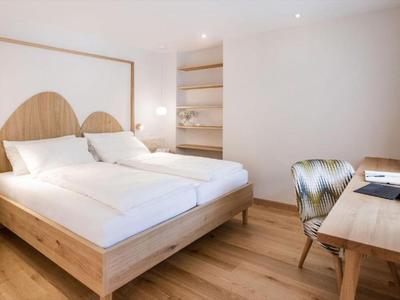 Helles Hotelzimmer mit Doppelbett, Holzmöbeln, Schreibtisch und Fenster mit Vorhängen.