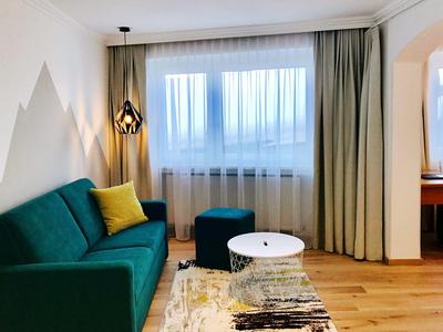 Habitación de hotel luminosa con sofá verde, cojín amarillo, mesa blanca pequeña y ventana con cortinas.