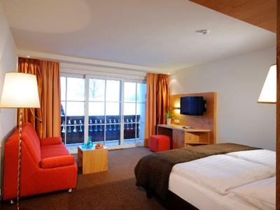 Habitación de hotel luminosa con cama doble, sofá rojo, televisor y ventana grande con balcón.