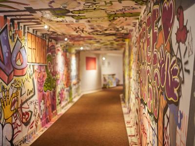Pasillo de hotel colorido con paredes con grafitis y suelo alfombrado que conduce a una habitación bien iluminada.
