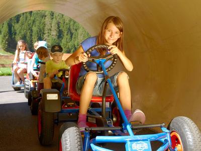 Children ride pedal go-karts through an outdoor tunnel on a sunny day.