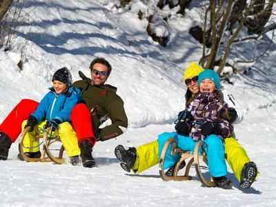 Zwei Paare sitzen auf Schlitten im Schnee, umgeben von verschneiter Landschaft und Bäumen.