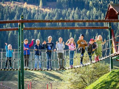 Children walk on a suspension bridge outdoors with forest and hills in the background.