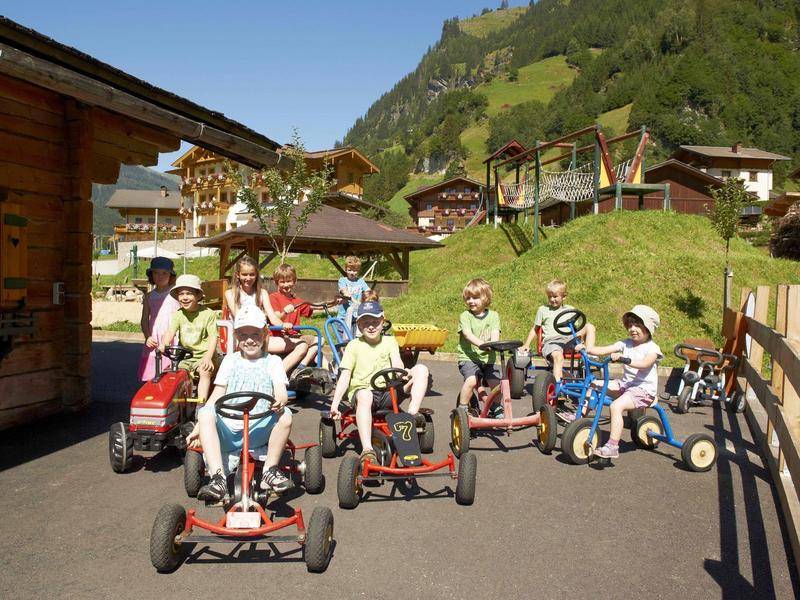 Gruppe von Kindern fährt mit Tretfahrzeugen vor einer Hütte in bergiger Landschaft.