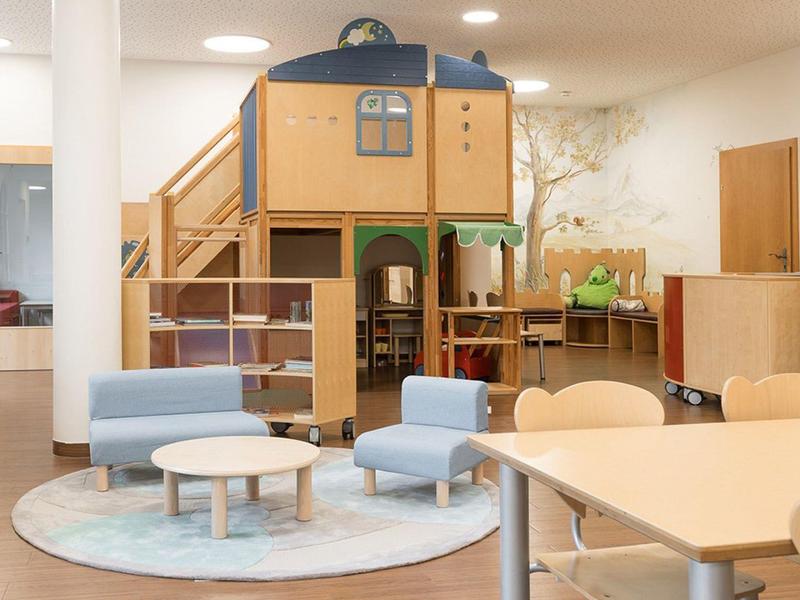 Kinderspielbereich mit Holzhäuschen, blauen Sesseln und kleinen Tischen in einem hellen Raum.