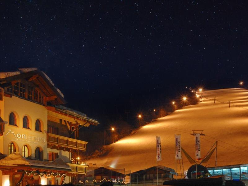 Nächtliche Skihangbeleuchtung und beleuchtetes Holzgebäude in verschneiter Berglandschaft.