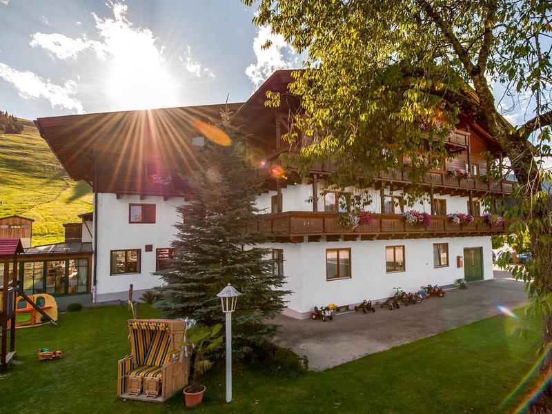 Großes weißes Haus mit Dachterrasse und Garten, Sonne strahlt durch Bäume, Hügel im Hintergrund.