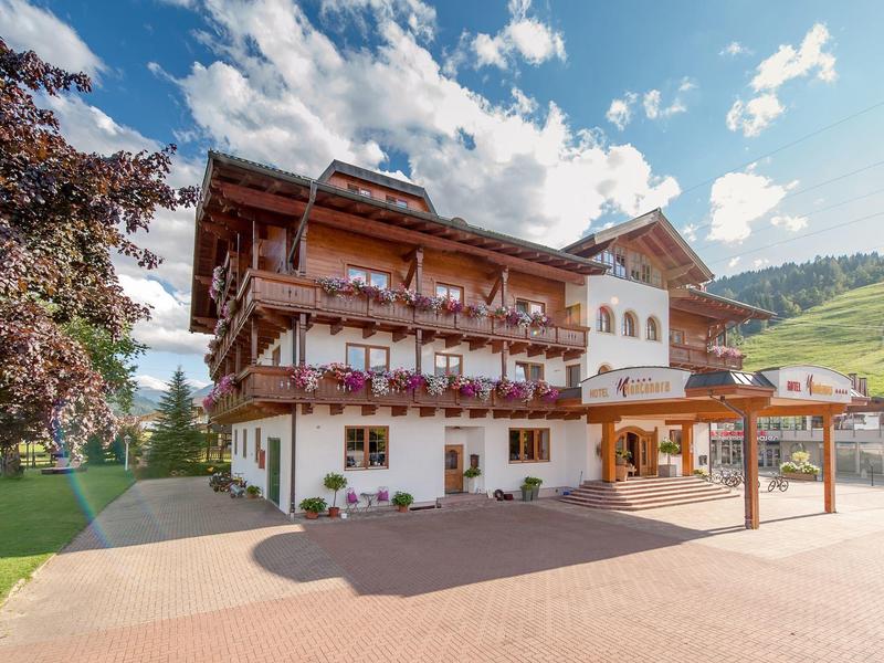 Großes Berghotel mit Holzbalkonen, Blumenschmuck, weißer Fassade und bewaldeter Hügellandschaft.