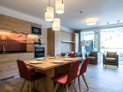 Salle à manger moderne avec table en bois et chaises rouges, adjacente à un salon lumineux avec grandes fenêtres.