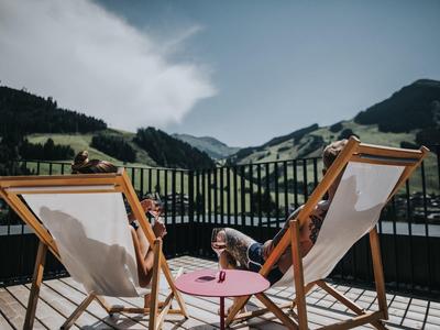 Deux personnes se détendent sur des chaises en bois avec vue sur un paysage montagneux sous un ciel nuageux.