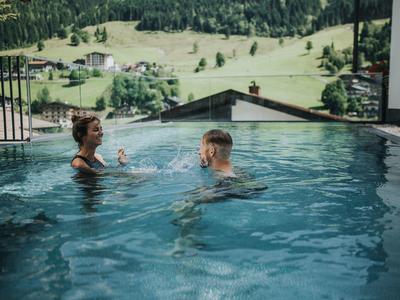 Deux personnes dans une piscine profitent de boissons avec vue sur des collines verdoyantes.