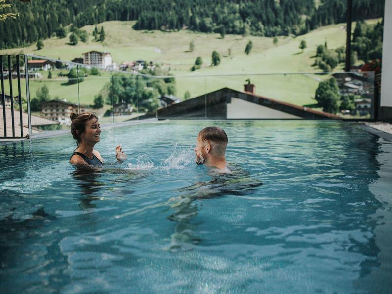 Deux personnes dans une piscine profitent de boissons avec vue sur des collines verdoyantes.