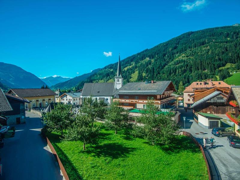 Kleines Dorf mit grüner Wiese, traditionellen Häusern und Bergen im Hintergrund unter blauem Himmel.