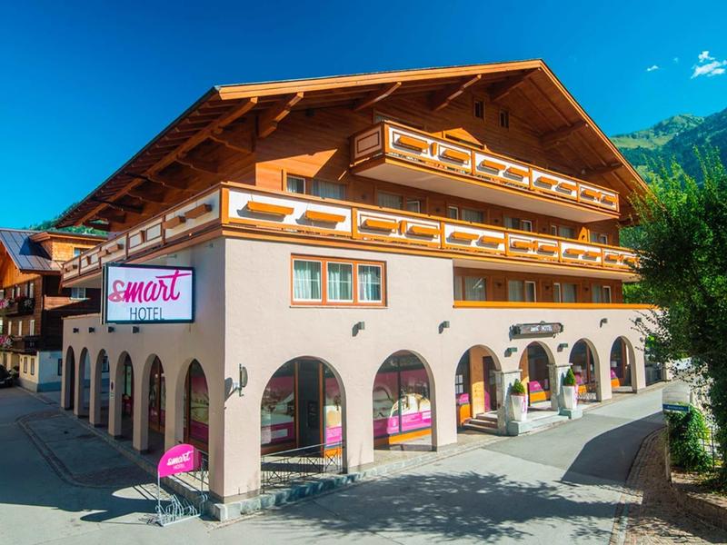 Großes, helles Chalet mit Holzbalkonen und rosa Schildern, umgeben von Bergen und klarem Himmel.