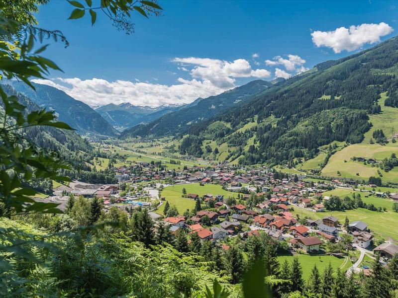 Blick auf ein grünes Tal mit Dorf, Wald und Bergen unter blauem Himmel und weißen Wolken.