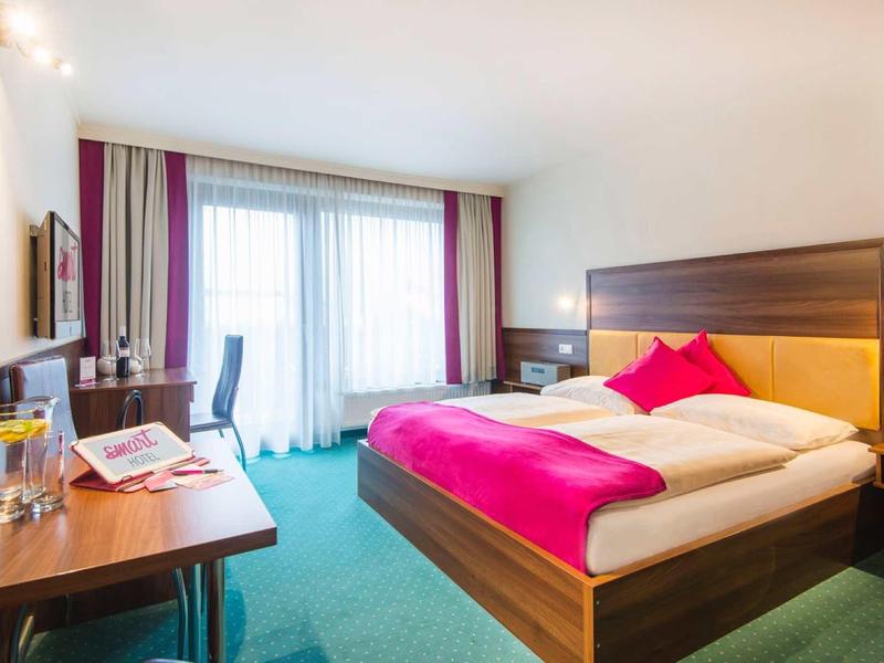 Helles Hotelzimmer mit Doppelbett, pinken Akzenten, Schreibtisch, Stuhl und großem Fenster mit Vorhängen.