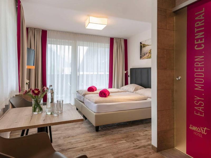 Helles, modernes Hotelzimmer mit Doppelbett, großem Fenster und Holzboden. Naturfarben und rote Akzente.