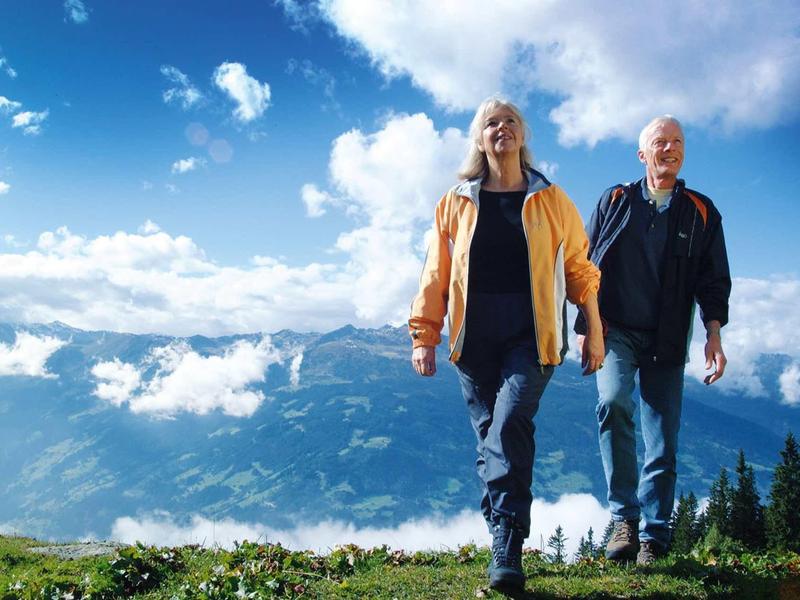 Couple âgé marchant dans un paysage de montagne sous ciel dégagé et soleil.