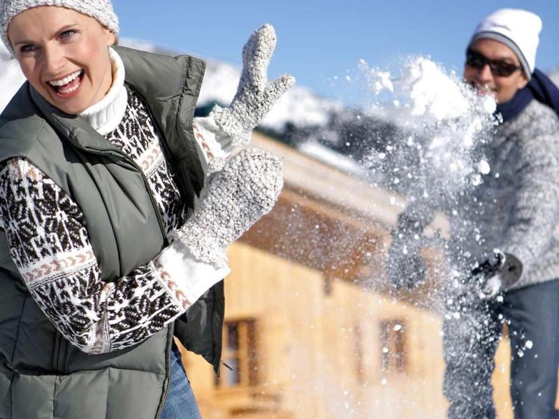 Deux personnes font une bataille de boules de neige devant une maison en bois dans la neige par une journée ensoleillée.