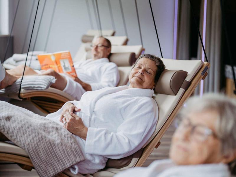 Trzy osoby w białych szlafrokach odpoczywają na wiszących leżakach w nowoczesnym wnętrzu.