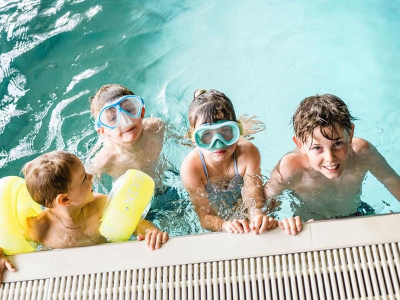 Cztery dzieci w basenie z goglami i rękawkami przy krawędzi basenu.