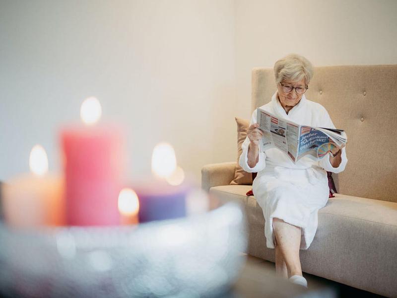 Starsza kobieta w szlafroku czyta na sofie przy świetle świec.