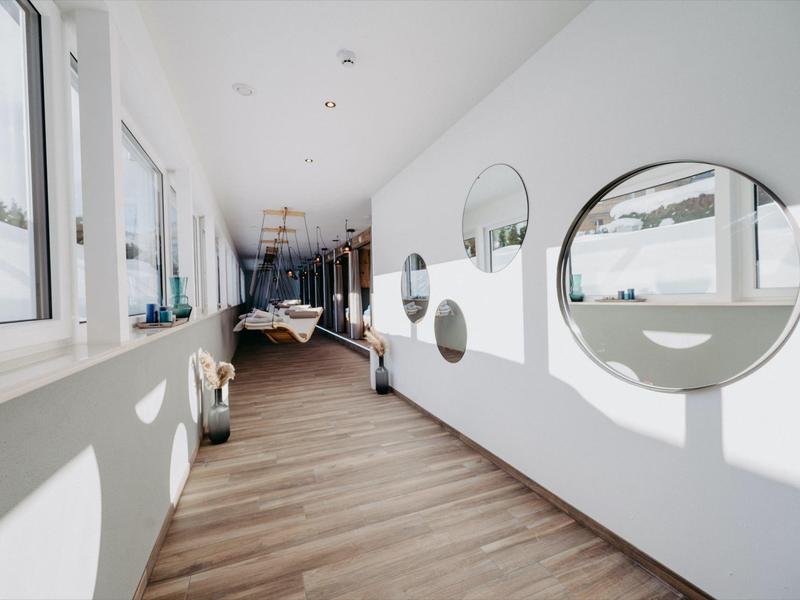 Długi jasny korytarz z drewnianą podłogą, okrągłymi lustrami na ścianie i oknami z widokiem na zaśnieżony krajobraz.