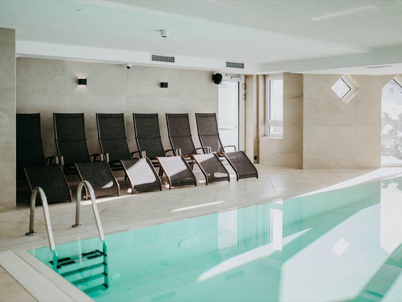 Basen kryty z leżakami i dużymi oknami w jasnej strefie wellness.