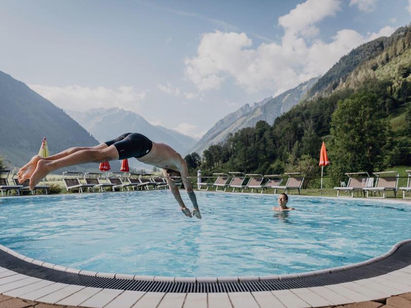 Osoba skáče do kulatého bazénu v horské krajině s lehátky.