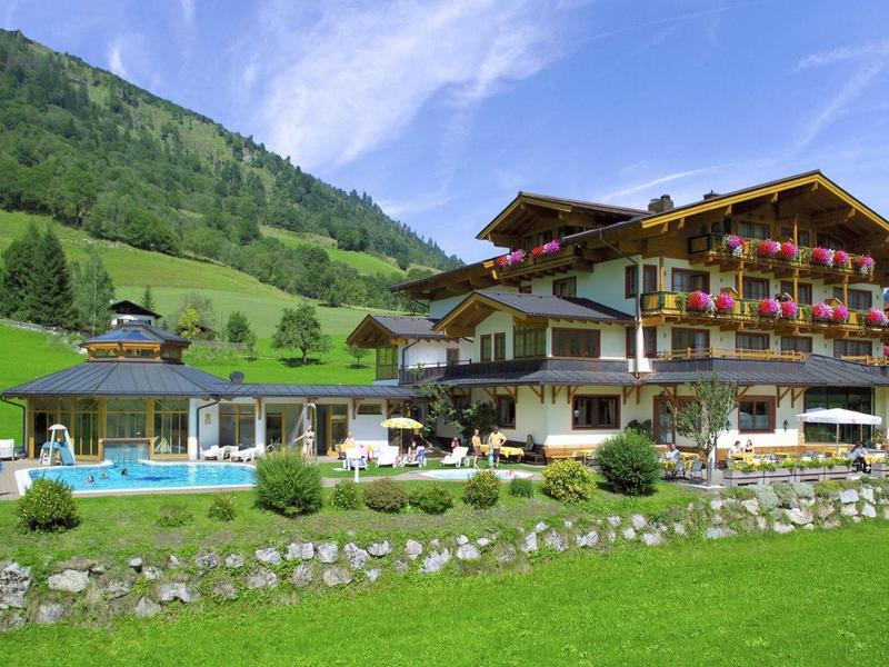 Alpský hotel s balkonem ozdobeným květinami a venkovním bazénem před zelenými kopci.