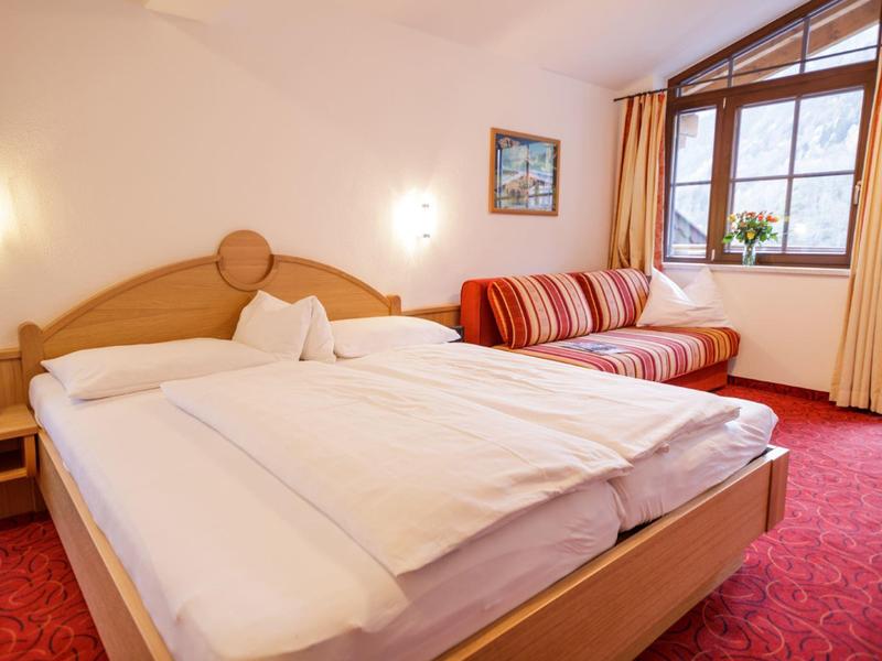 Pohodlný hotelový pokoj s manželskou postelí, červeným kobercem a pohovkou u okna.