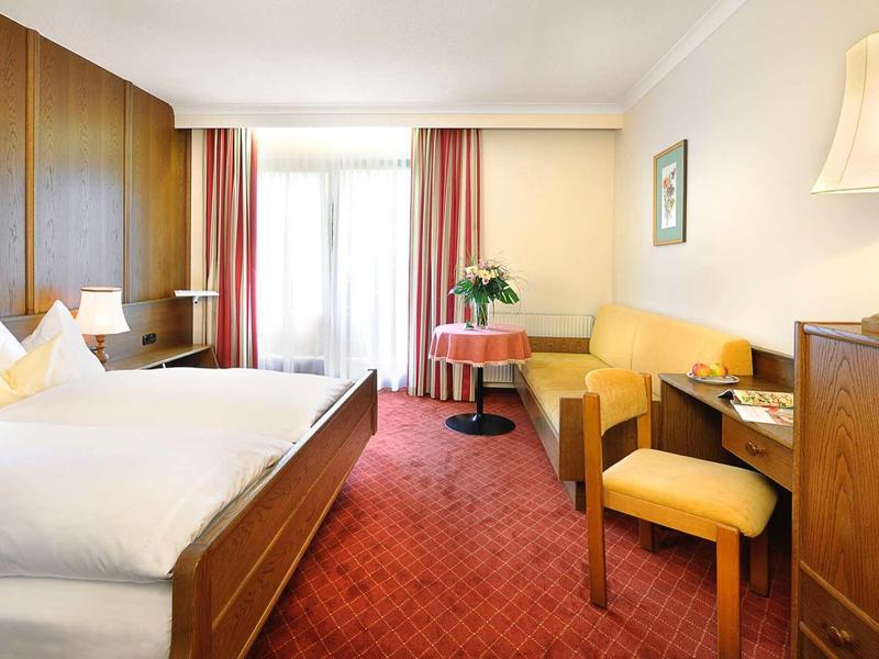 Jasny pokój hotelowy z łóżkiem podwójnym, czerwonym dywanem, biurkiem, krzesłem i zasłonami przy oknie.