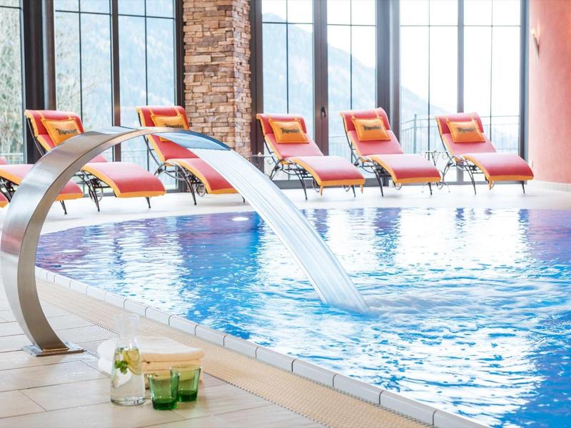 Piscine intérieure avec fontaine d'eau et chaises longues orange devant de grandes fenêtres.