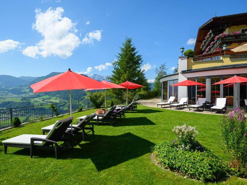 Chaises longues avec parasols rouges sur pelouse verte devant un hôtel avec vue sur la montagne.