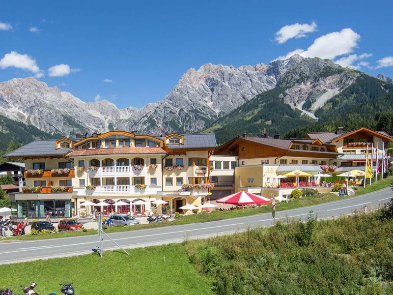 Grand hôtel avec décor alpin, prairies vertes et ciel bleu.