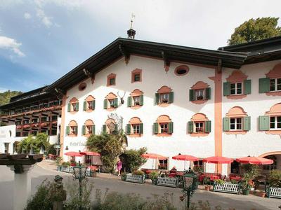 Großes weißes Haus mit roten Fensterläden, Gartentischen und grünen Bäumen im Hintergrund.