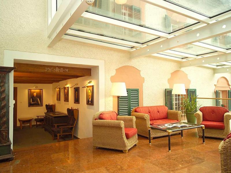 Světlý lounge s pohovkami, křesly a velkými okny pod skleněnou střechou.